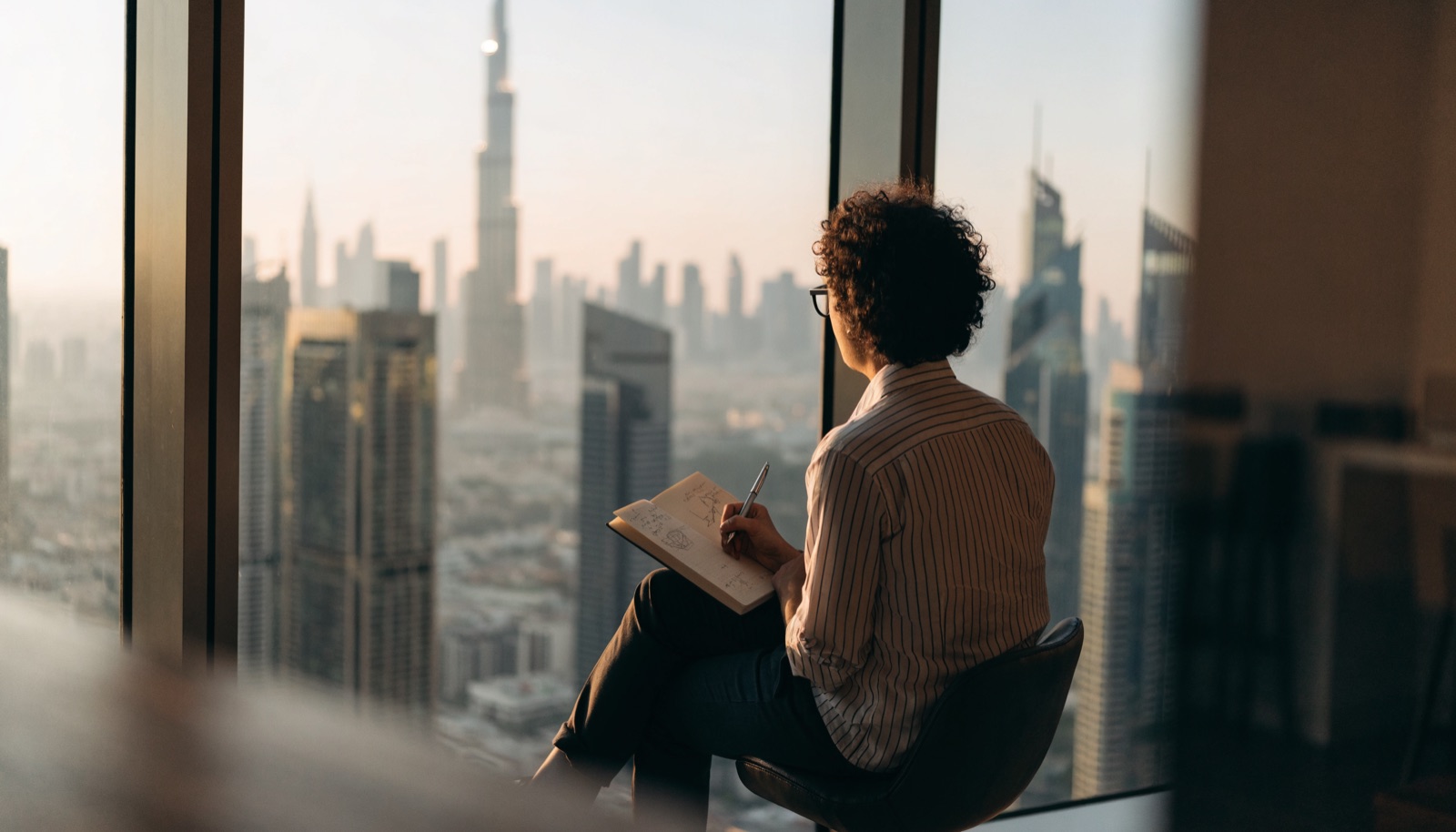 A founder sketching plans at a window overlooking the Dubai skyline at dawn
